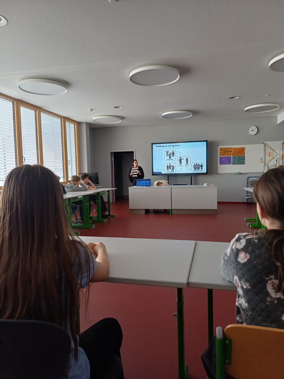 A classroom with students seated at tables. A woman stands in front of a projector screen, likely presenting. The room has large windows and round ceiling lights.