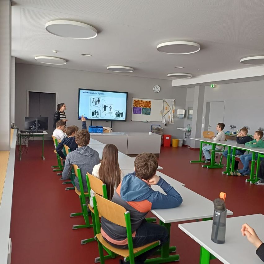 In a classroom, a teacher stands in front of a projector screen showing a diagram. Students sit at tables, listening attentively. The room has red flooring and ceiling lamps.