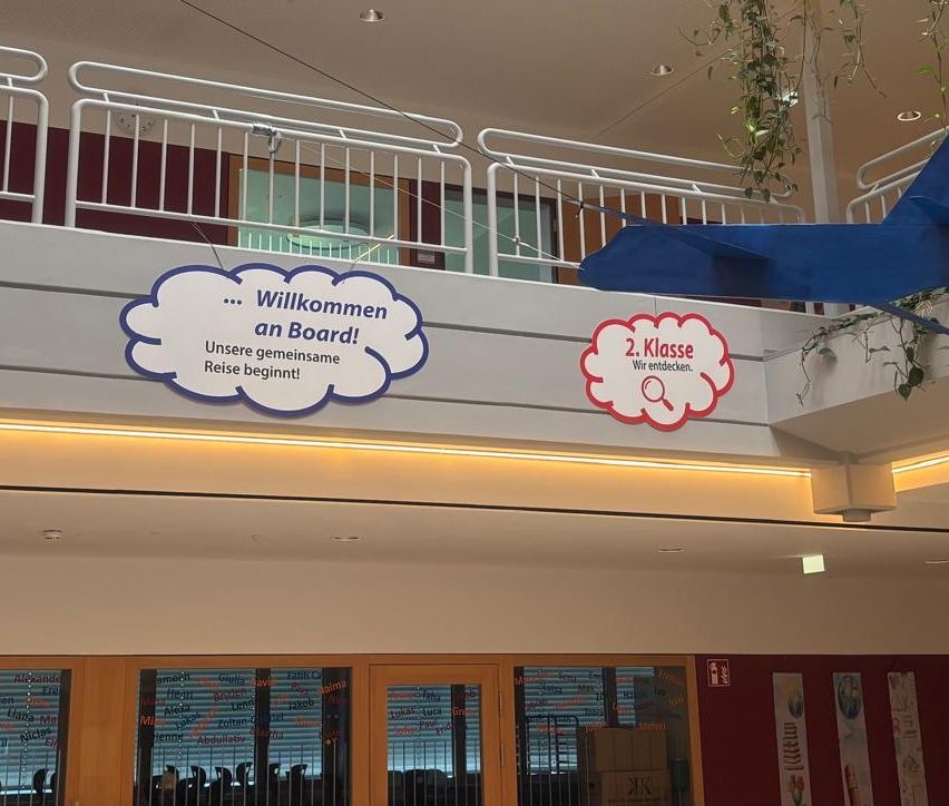 An indoor area with a white railing and two signs. One sign says 'Willkommen an Board!' and the other says '2. Klasse Wir entdecken.' There are also plants hanging from the ceiling.