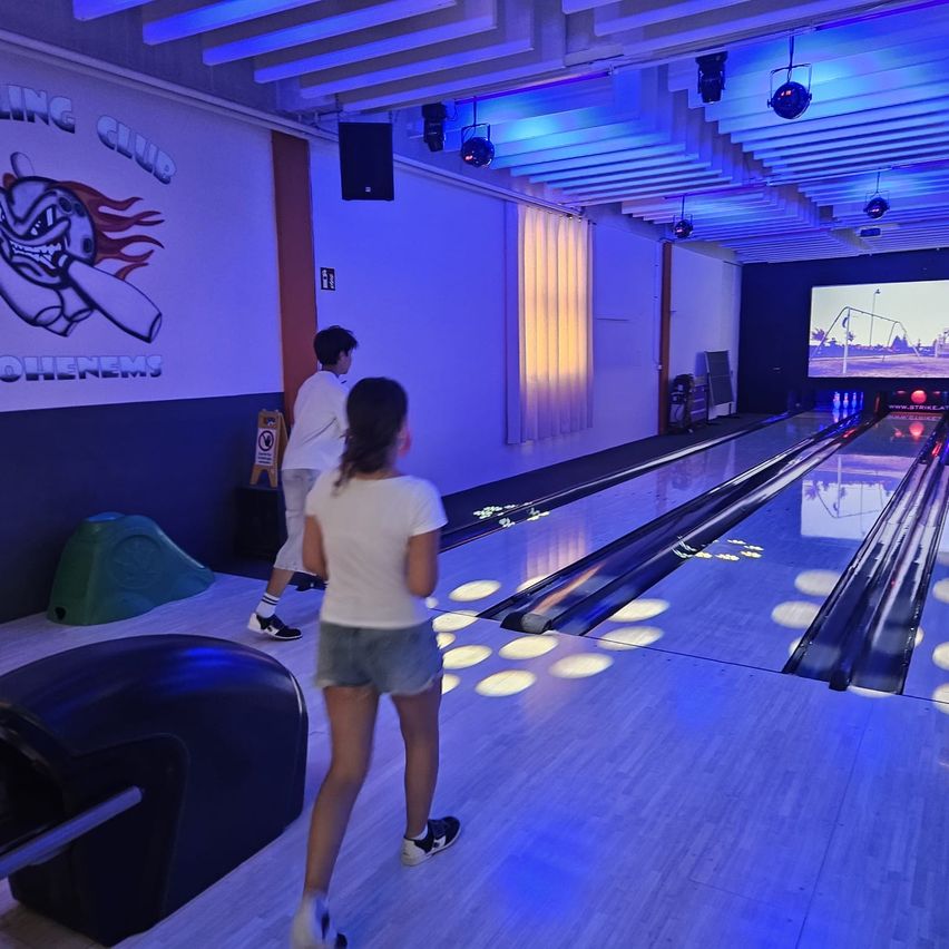 Zwei Personen spielen Bowling in einer hell beleuchteten Bowlingbahn mit blauer Beleuchtung und einer großen Leinwand. Eine Person steht bereit zum Werfen, während eine andere wartet.