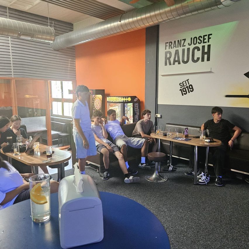 Eine Gruppe von Menschen sitzt auf einer Couch in einem Raum mit orangefarbenen Wänden. Ein Mann steht vor ihnen. Der Raum hat einen Tisch mit Getränken und einem Dartboard.