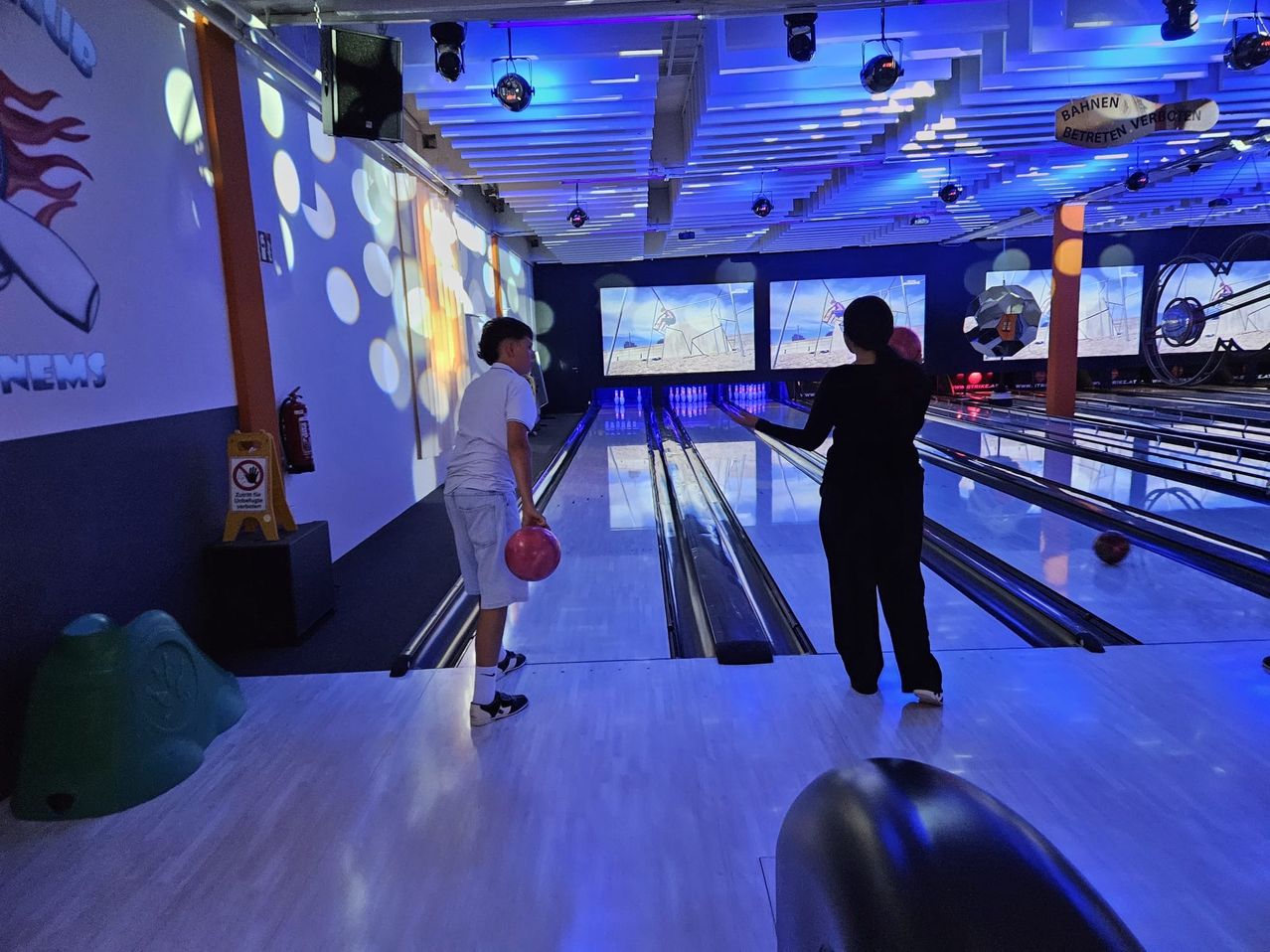 Ein Mann und eine Frau in einer Bowlingbahn. Der Mann hält einen Bowlingball. Die Frau steht an der Bahn.