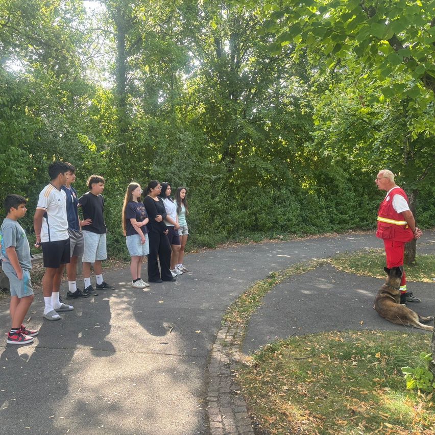Eine Gruppe junger Menschen steht in einer Reihe auf einem Pfad in einem Park. Ein Mann in einer reflektierenden Weste steht in der Nähe, und ein Hund sitzt neben ihm.