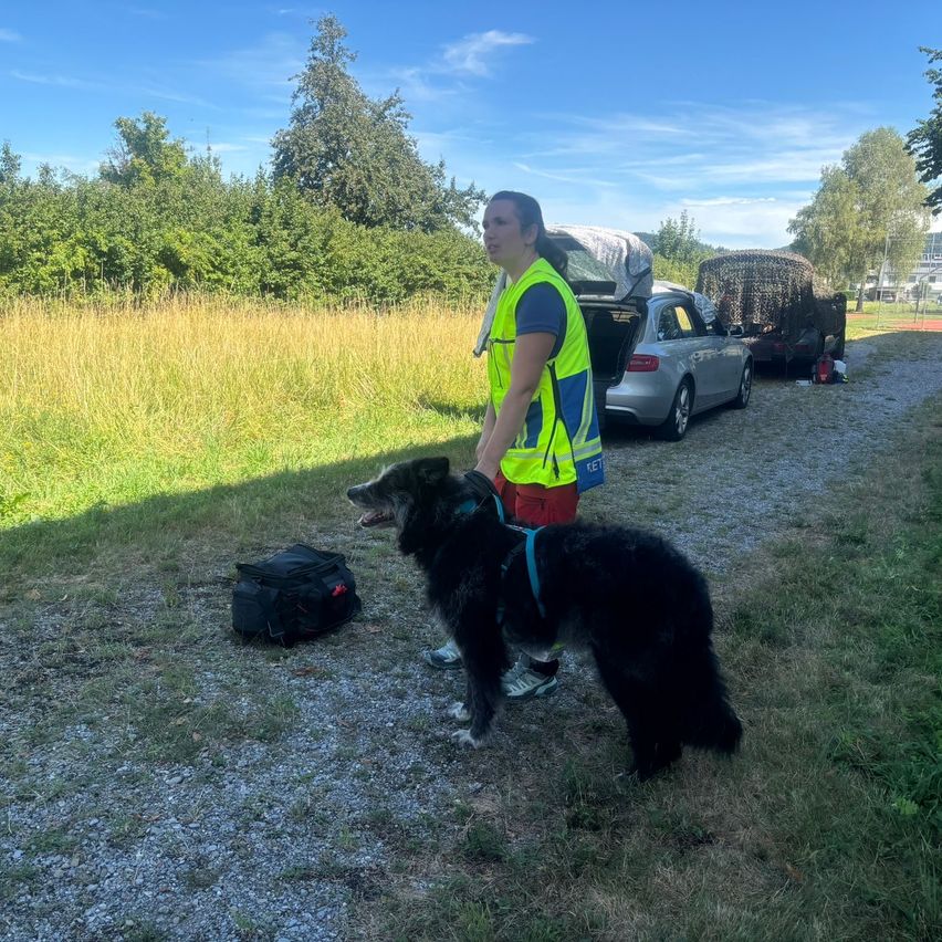 Eine Frau in einer gelben Weste und roten Hosen führt ihren großen schwarzen Hund mit einem blauen Halsband auf einem Kiesweg neben einem Auto und einer Tasche.