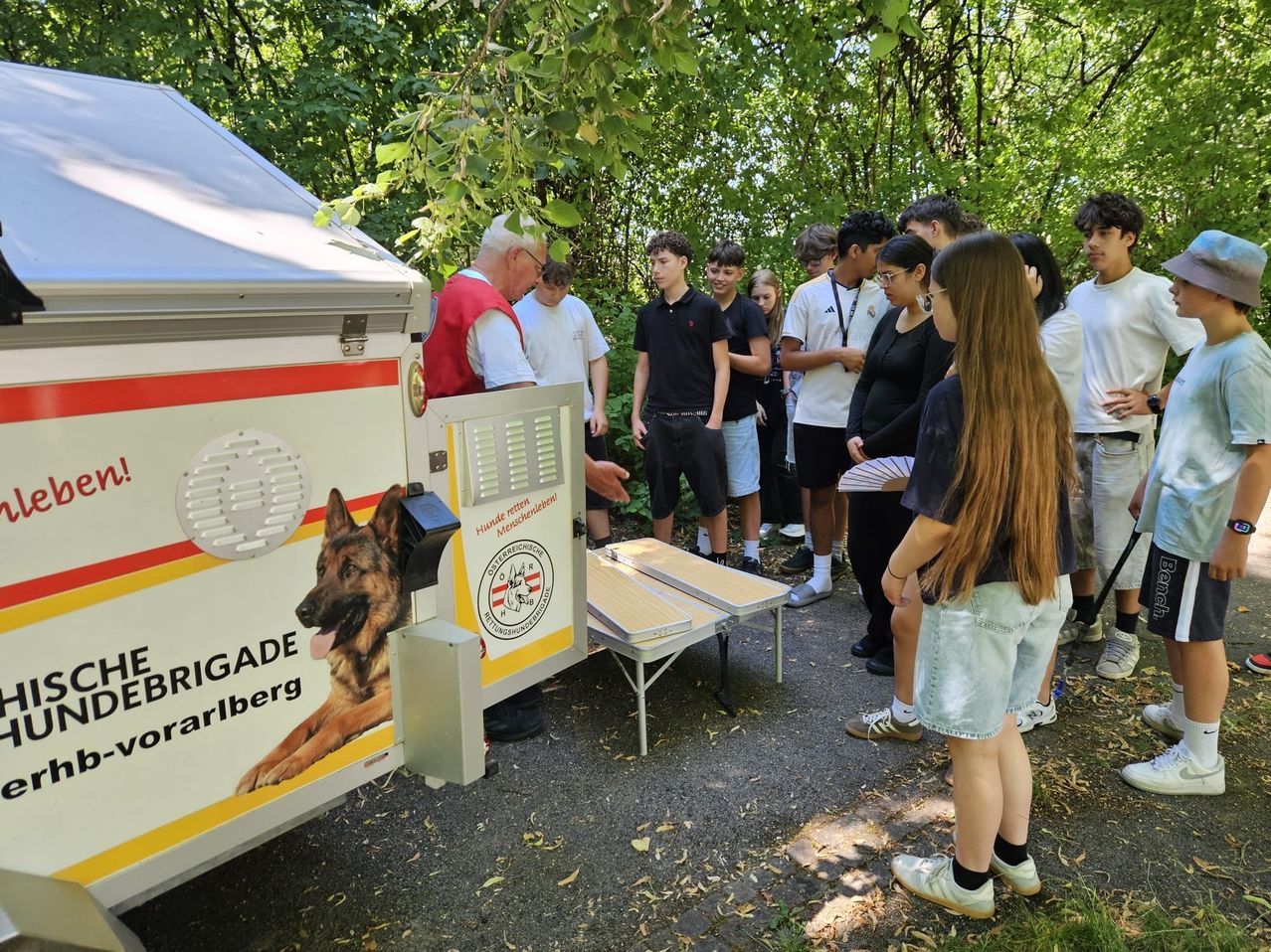 Eine Gruppe von Schülern und ein Mann versammeln sich um eine mobile Ausbildungseinheit. Die Einheit zeigt einen deutschen Schäferhund und die Worte 'Ostereinigung'. Die Schüler hören aufmerksam dem Mann zu.