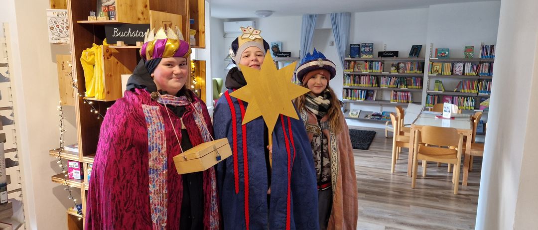 Drei Personen in festlichen Kostümen stehen in einem Raum mit Holzboden. Einer hält einen Stern, ein anderer eine Kiste und alle tragen Kronen. Hinter ihnen sind mit Büchern gefüllte Regale zu sehen.