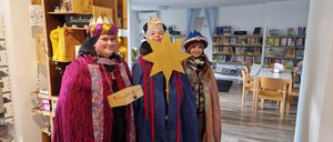 Drei Personen in festlichen Kostümen stehen in einem Raum mit Holzboden. Einer hält einen Stern, ein anderer eine Kiste und alle tragen Kronen. Hinter ihnen sind mit Büchern gefüllte Regale zu sehen.