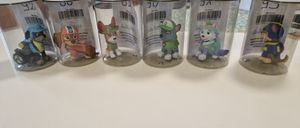 Fünf durchsichtige Behälter mit weißen Deckeln zeigen kleine Figuren von Paw Patrol Charakteren. Jeder Behälter hat einen Strichcode. Im Hintergrund ist ein Regal mit Büchern und einer Tasse zu sehen.