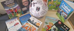 Ein signierter Fußballball steht zwischen Büchern, darunter ein Buch über Schatten, ein Buch von James von Leyden und ein Kinderbuch über Fußball.