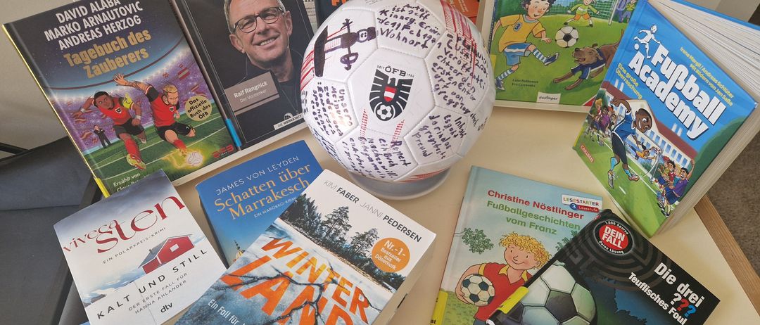 Ein signierter Fußballball steht zwischen Büchern, darunter ein Buch über Schatten, ein Buch von James von Leyden und ein Kinderbuch über Fußball.