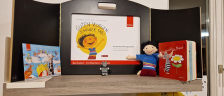 A book display with a cartoon character on the cover. The title reads 'Guten Morgen! Schöner Tag!' Two stuffed toys are on the shelf below.