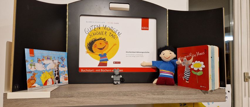 Ein Buchdisplay mit einem Cartoon-Charakter auf dem Cover. Der Titel lautet 'Guten Morgen! Schöner Tag!' Zwei Plüschtiere stehen auf dem Regal darunter.