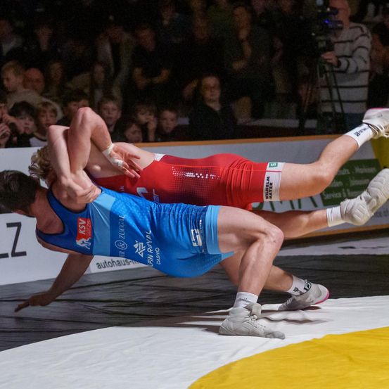 Zwei Ringer sind in einem Kampf auf einer Matte in einer Sportarena. Einer trägt ein blaues Trikot, der andere ist in Rot. Zuschauer beobachten den Kampf.