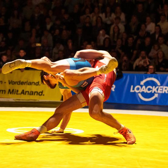 Zwei Ringer in blauen und roten Outfits kämpfen auf dem Ring. Ein Ringer ist in der Luft, während der andere am Boden liegt. Hinter ihnen befindet sich eine gelbe Barriere mit einer blauen Werbung.
