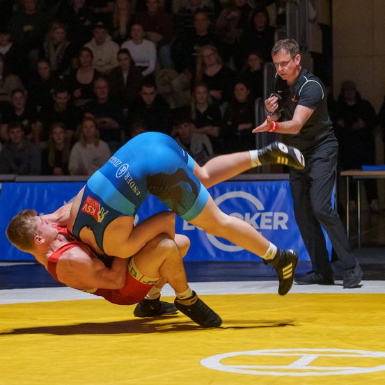 Zwei Ringer kämpfen auf einem gelben Teppich vor einem Publikum. Ein Ringer trägt Blau und der andere Rot. Ein Schiedsrichter beobachtet in der Nähe.