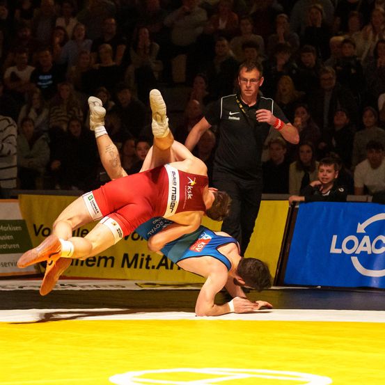 Zwei Ringer in Rot und Blau sind mitten in einer Aktion auf einem gelben Mattenring, einer in der Luft und der andere am Boden. Ein Schiedsrichter beobachtet aufmerksam.