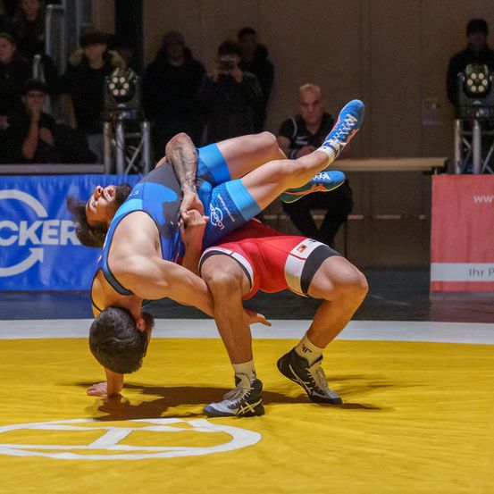 Zwei Männer kämpfen in einem Wettkampf auf einem gelben Mattenring. Einer trägt ein blaues Trikot, der andere ein rotes. Sie halten sich gegenseitig in einer Ringposition fest. Im Hintergrund schauen Zuschauer zu.