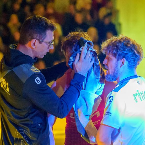 Ein Mann mit Brille justiert das Handtuch um den Hals eines anderen Mannes vor einer jubelnden Menge.