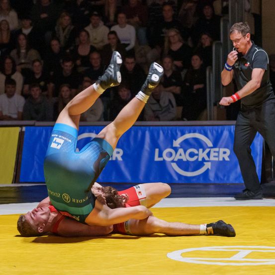 Zwei Ringer in blauen und roten Uniformen befinden sich mitten in einem Ringkampf. Einer ist oben, der andere unten. Ein Offizieller steht in der Nähe. Das Publikum schaut von den Tribünen zu.