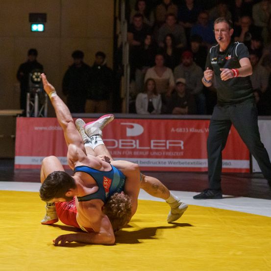 Ein Wrestling-Match findet auf einer gelben Matte statt. Ein Schiedsrichter beobachtet von der Seite, während Zuschauer im Hintergrund sitzen. Ein Wrestler hält den anderen in einem Griff.