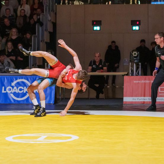 Zwei Ringer in einer Turnhalle führen eine Grifftechnik vor einem Publikum durch. Ein Offizieller steht in der Nähe und beobachtet den Kampf.