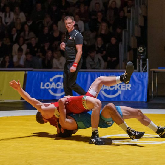 Zwei Ringer befinden sich in einem Ringkampf auf einem gelben Teppich, während ein Schiedsrichter sie beobachtet. Das Publikum sitzt hinter einem blauen Banner.