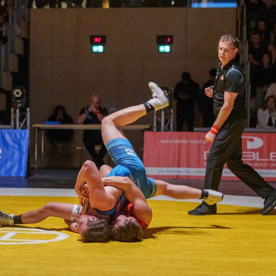 Zwei Ringer in blauen und roten Uniformen kämpfen auf einem gelben Teppich. Ein Schiedsrichter in Schwarz steht in der Nähe. Das Publikum sitzt im Hintergrund und beobachtet das Match.