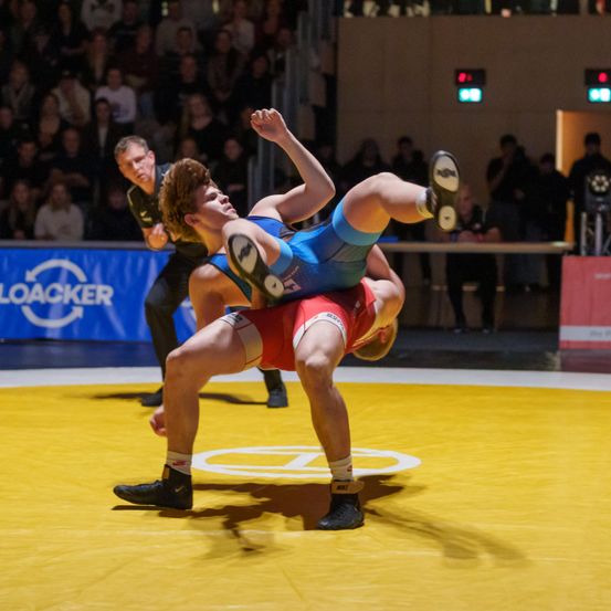 Zwei männliche Ringer in blauen und roten Singlets sind in Aktion, einer der Ringer ist hochgehoben. Der Schiedsrichter steht hinter ihnen, und Zuschauer beobachten von den Tribünen aus.