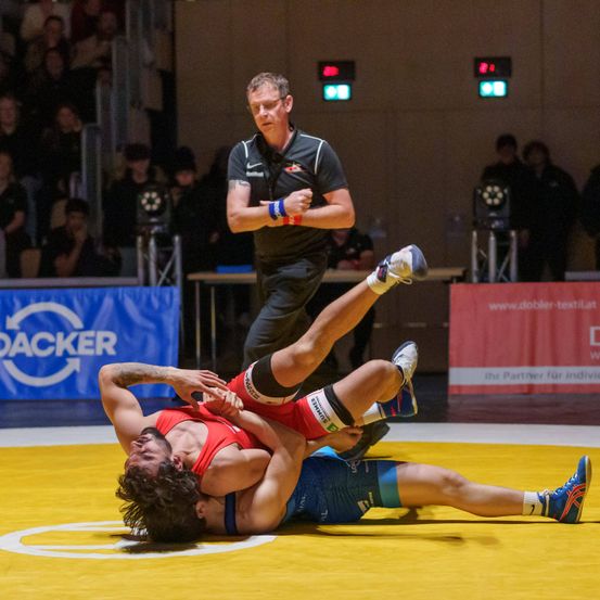 Zwei Ringer kämpfen auf einem gelben Teppich, einer oben. Ein Schiedsrichter steht hinter ihnen. Ein Publikum schaut im Hintergrund zu.