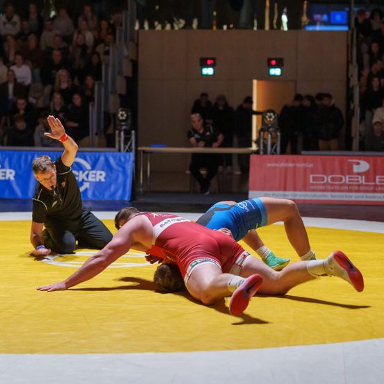 Zwei Männer ringen auf einem gelben Teppich. Einer trägt ein rotes Trikot, der andere ist in Blau. Ein Schiedsrichter signalisiert hinter ihnen. Das Publikum schaut von den Tribünen zu.