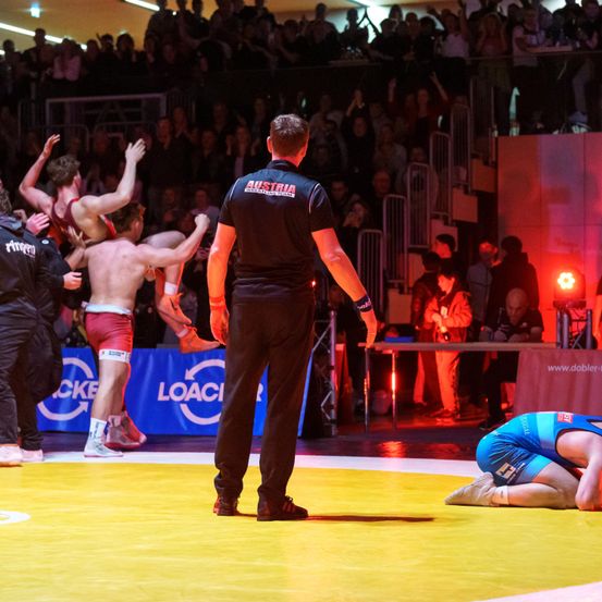 Zwei Ringer kämpfen in einem Ring, einer auf den Knien, während ein Schiedsrichter zusieht. Zuschauer in den Rängen jubeln, und ein Scheinwerfer leuchtet von hinten.