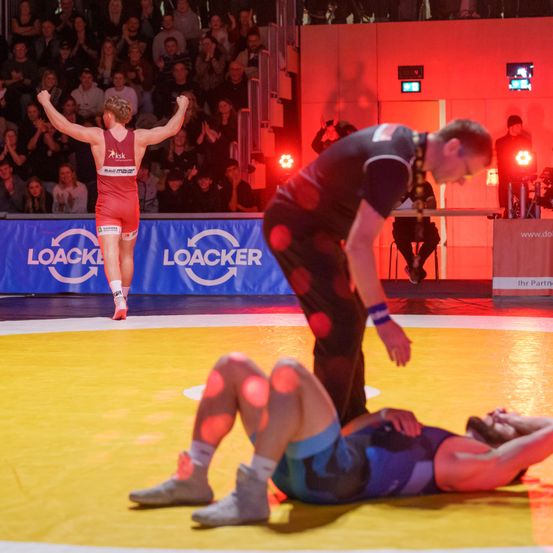 Ein Ringer in einem roten Singlet feiert einen Sieg auf einem gelben Matten, mit einem Schiedsrichter in der Nähe und Zuschauern im Hintergrund.