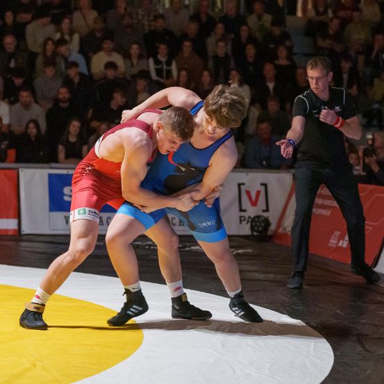 Zwei junge Männer ringen auf einem Teppich, einer trägt ein rotes Trikot und der andere Blau. Ein Schiedsrichter steht in der Nähe und beobachtet sie. Das Publikum sitzt im Hintergrund, einige mit ihren Telefonen in der Hand.