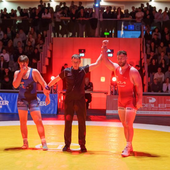 Drei Ringer stehen auf einem Ring. Der Schiedsrichter hebt die Hand des Siegers. Das Publikum sitzt auf den Tribünen.