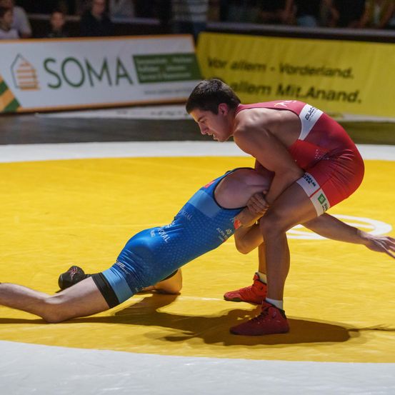 Zwei Wrestler treten in einem Match auf einem gelben Teppich an. Der Mann in Blau liegt auf dem Teppich, während der Mann in Rot ihn fixiert. Sie tragen beide rote Turnschuhe.