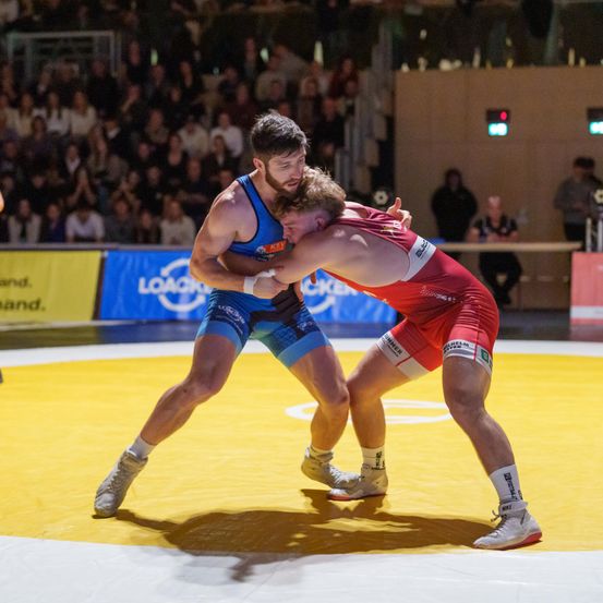 Zwei Ringer in blauen und roten Uniformen sind in einem Kampf auf einem gelben Mattenring engagiert. Ein Publikum sitzt im Hintergrund.