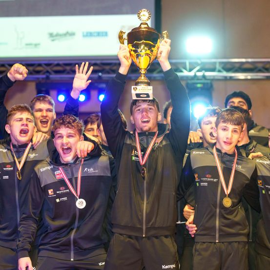 Eine Gruppe junger Männer in schwarzen Uniformen feiert, einer hält einen goldenen Pokal mit einer Schleife. Sie stehen in einer Reihe, lächeln und heben ihre Hände.