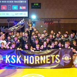 Eine Sportmannschaft feiert ihren Sieg mit einer Trophäe und einem Banner, das KSK Hornets zeigt. Sie tragen alle Medaillen und halten Fahnen. Dahinter steht eine große Anzeigetafel mit einer Anzeigetafel.