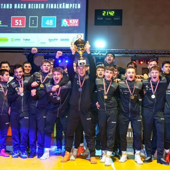 Eine Gruppe von Athleten mit Medaillen und einem Pokal posiert für ein Foto auf der Bühne. Der Hintergrund zeigt eine Anzeigetafel mit Punkten.