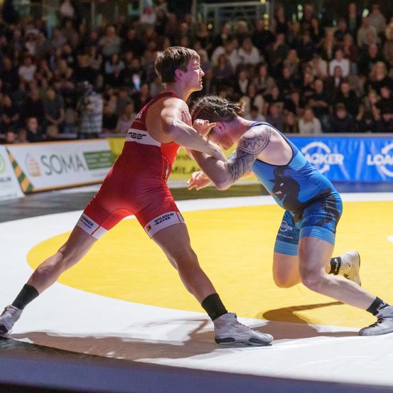Zwei Ringer in einem Wettkampf, einer in Rot und der andere in Blau, ringen intensiv auf einer Ringermatte, während eine Menge zuschaut.