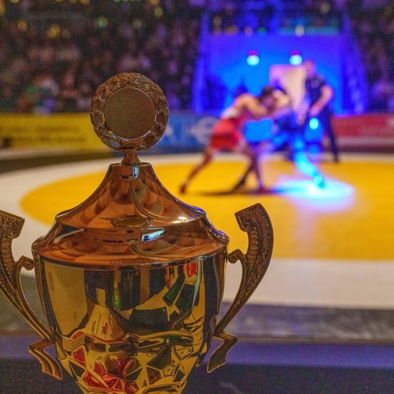 Ein Pokal im Vordergrund mit zwei ringen auf einem gelben Teppich im Hintergrund. Das Publikum ist verschwommen.