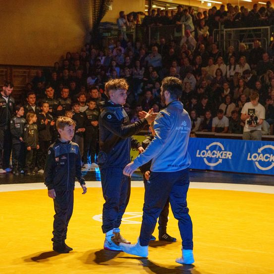 Zwei Männer schütteln sich in der Mitte eines Wrestlingrings die Hände, ein junger Junge steht zwischen ihnen, eine Menge beobachtet.