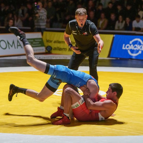 Zwei Männer ringen auf einem gelben Teppich, einer in Blau und der andere in Rot. Ein Schiedsrichter steht in der Nähe und beobachtet genau. Im Hintergrund sieht ein Publikum das Match.