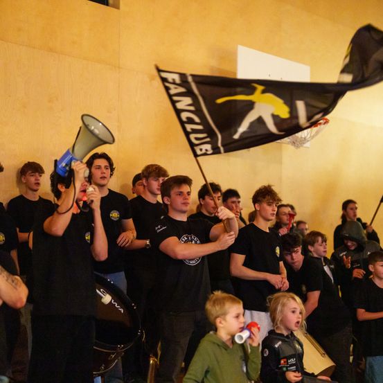 Eine Gruppe von Menschen in schwarzen T-Shirts steht in einem Raum, einige halten eine Fahne und andere benutzen Megaphone. Ein Junge und ein Mädchen vorne scheinen aufgeregt zu sein. Ein Trommel steht links.