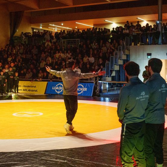 Ein Mann in einer grauen Jacke geht mit ausgestreckten Armen über einen gelben Ring. Drei Männer in grünen Uniformen stehen in der Nähe. Eine große Menge sieht von den Tribünen zu. Banner sind sichtbar, darunter eines mit der Aufschrift LOACKER.