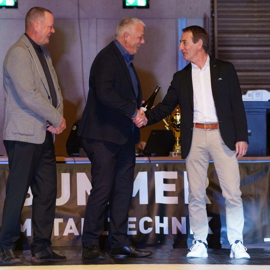 Drei Männer stehen auf einer Bühne, zwei schütteln sich die Hände, während der dritte eine Flasche hält. Dahinter steht ein Tisch mit einem goldenen Pokal und einem Banner, auf dem 'Summer Metal Tech' steht.