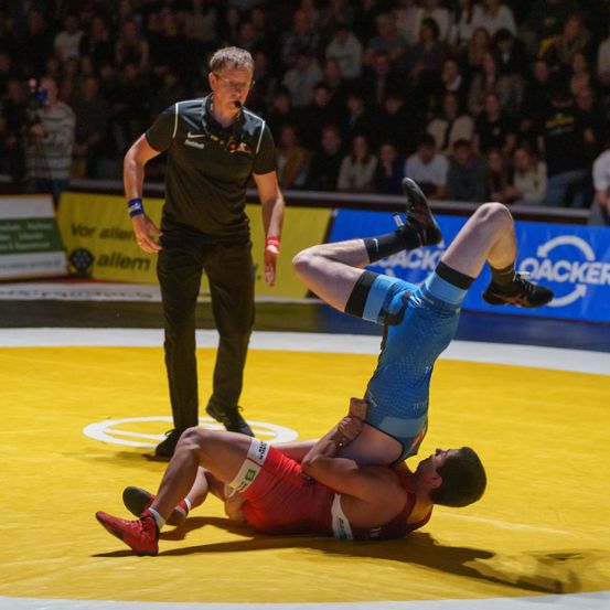 Zwei Ringer kämpfen auf einer gelben Matte, einer auf dem anderen. Ein Offizieller steht hinter ihnen. Das Publikum beobachtet vom Publikumsrang.