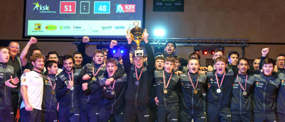 Ein Sportteam mit Medaillen feiert mit einem Pokal. Die Anzeigetafel hinter ihnen zeigt ein Ergebnis von 51 zu 48. Das Team trägt passende schwarze und graue Uniformen.