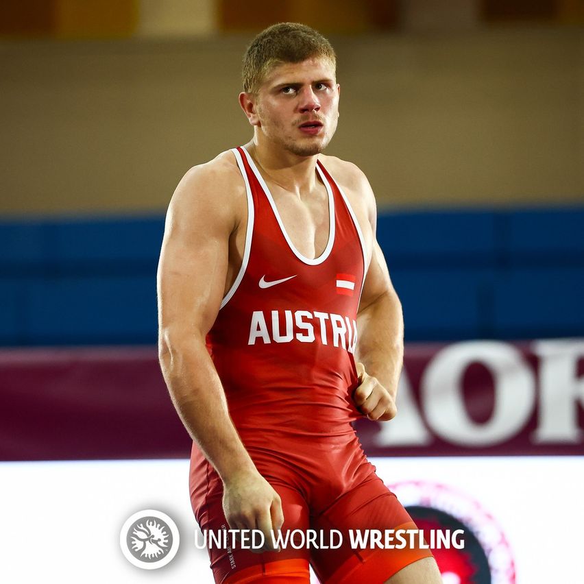 Ein Ringer in roter Kleidung, die Österreich repräsentiert, läuft auf dem Teppich. Auf seinem Shirt steht 'AUSTR', und unten steht 'United World Wrestling'.