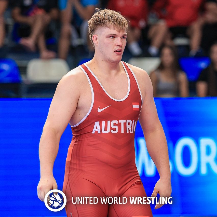 Ein Ringer in einem roten Trikot mit Austria- und Nike-Logos steht vor einem blauen Hintergrund mit dem United World Wrestling Logo.